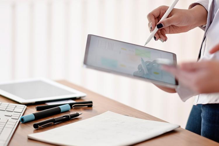 Imagen donde se ve una mano sujetando un lápiz táctil enfrente de una tableta. Debajo de ella hay un escritorio lleno de papeles y material de escritura.