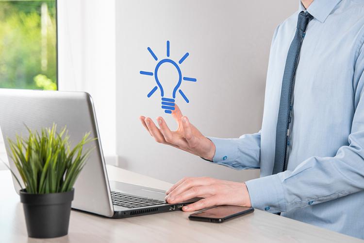 Hombre con camisa y corbata sostiene en su mano derecha una bombilla azul pintada digitalmente. La otra mano está apoyada en un portátil, el cual tiene una planta detrás.