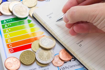 Una mano con un boli apunta información en una agenda. En la mesa aparecen monedas de diferente valor sobre una hoja con el gráfico de los diferentes tipos de eficiencia energética (A, B, C...)